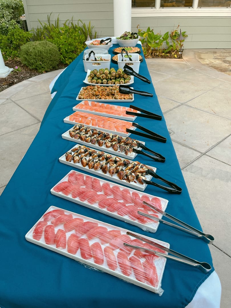 A long table with a blue tablecloth holds various sushi and appetizer platters outdoors.