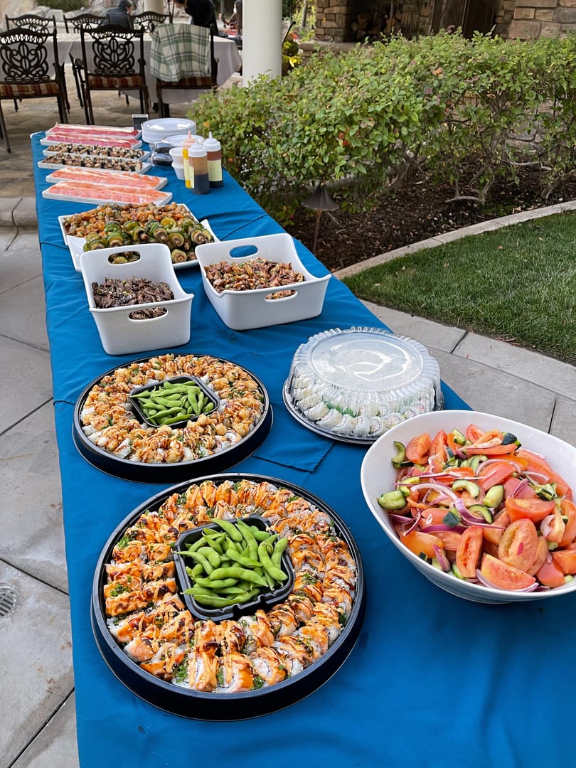 A blue-covered table laden with various sushi platters, salads, and other dishes at an outdoor event.