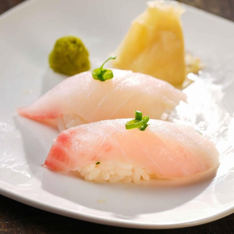Two pieces of nigiri sushi with light pink fish, green onions, wasabi, and ginger.