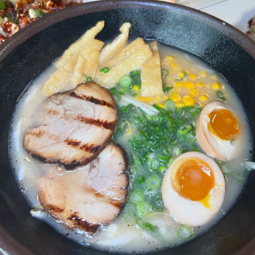 Close-up of a ramen bowl with pork, soft-boiled eggs, corn, green onions, and fried tofu.