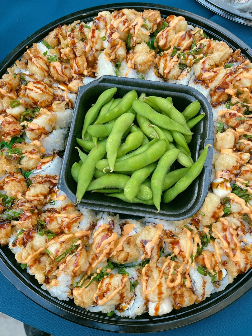 A platter of sushi rolls topped with fried shrimp, sauces, and green onions, with a side of edamame.
