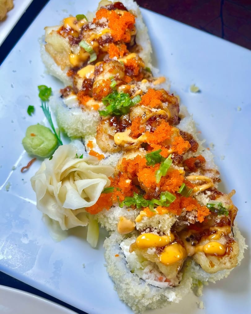 Sushi rolls topped with fried shrimp, orange roe, green onions, and various sauces on a white plate.