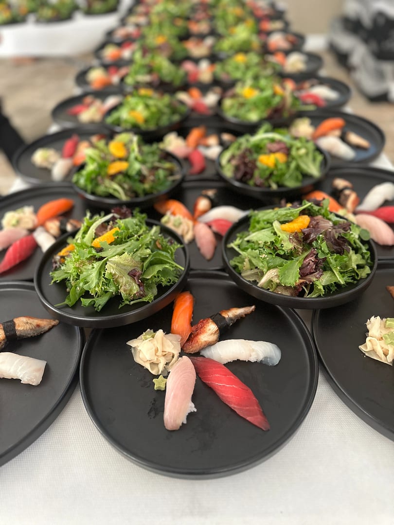 Rows of black plates with sushi and side salads, set for a large gathering.