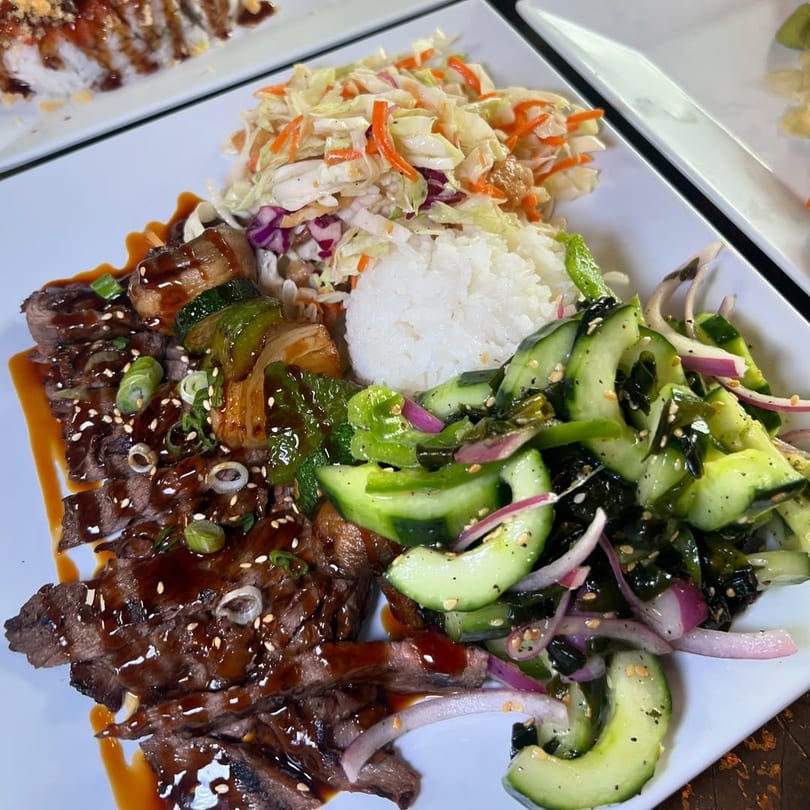 Grilled steak with teriyaki sauce, rice, coleslaw, and cucumber salad on a white plate.